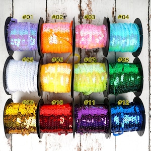 May include: Twelve spools of iridescent sequin trim in various colours, including purple, orange, pink, blue, white, yellow, green, red, gold, and blue. Each spool is labelled with a number from 01 to 12.