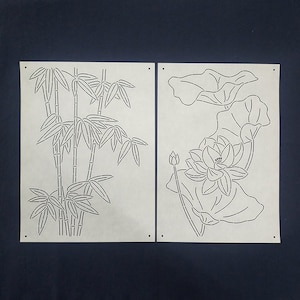 Può includere: Due stencil di carta bianca, uno con un disegno di bambù e l'altro con un disegno di fiore di loto. Entrambi gli stencil hanno un motivo a punti.