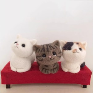 May include: Three felt cat figurines in white, brown tabby, and calico patterns sit on a small red platform. Each cat has large, dark eyes and a smiling expression, showcasing a whimsical, handmade aesthetic.