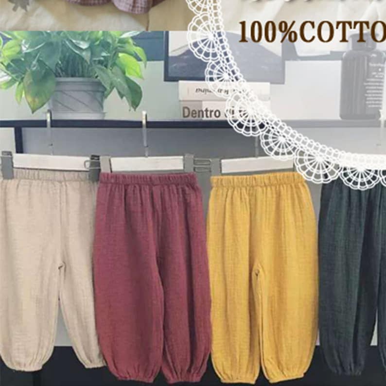 May include: Four pairs of children's trousers in beige, burgundy, yellow, and dark green, hanging on a rack. The trousers are made of a lightweight, textured fabric. The text "100% COTTON" is visible in the background.