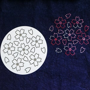 May include: A white circle stencil with a design of cherry blossoms and leaves. The stencil is on a dark blue fabric background with a similar design embroidered in pink and white thread.