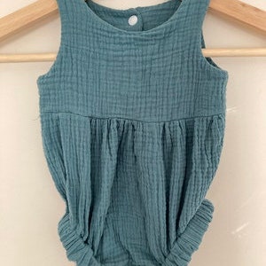 May include: A teal baby romper made from textured fabric, hanging on a wooden hanger. The romper has a sleeveless design, a gathered waist, and elasticated leg openings. A single button fastens at the back.