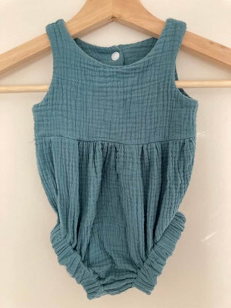 May include: A teal baby romper made from textured fabric, hanging on a wooden hanger. The romper has a sleeveless design, a gathered waist, and elasticated leg openings. A single button fastens at the back.