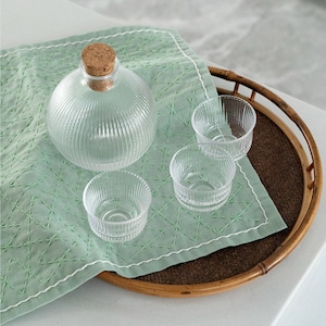 Puede incluir: Una botella de vidrio transparente con tapón de corcho y tres cuencos pequeños de vidrio transparente a juego. La botella y los cuencos tienen un diseño acanalado. Se encuentran sobre un paño verde claro con costuras blancas, dentro de una bandeja redonda de bambú.