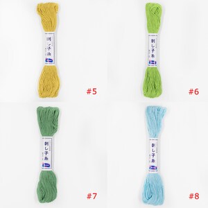 May include: Four skeins of embroidery floss in different colours: yellow, green, light green, and light blue. Each skein has a label with Japanese text that reads "刺し子糸" which translates to "Sashiko thread". The numbers 5, 6, 7, and 8 are written below each skein.