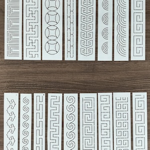 May include: A collection of sixteen white rectangular stencils featuring various geometric patterns, including Greek keys, interlocking circles, waves, and linear borders. Each stencil is designed with cut-out shapes for painting.