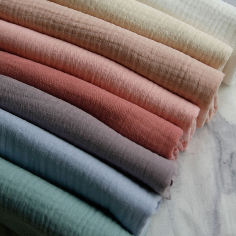 May include: A selection of folded fabric in a range of pastel hues, including pinks, beiges, blues, and greys. The fabric has a textured, crinkled look, indicating a soft, lightweight material. The arrangement highlights the variety of colours and textures.