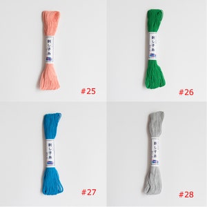 May include: Four spools of embroidery floss in different colours: peach, green, blue, and grey. Each spool has a white label with Japanese text and the number 25, 26, 27, or 28.