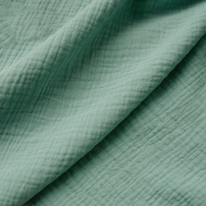 May include: A close-up shot of a folded piece of soft, textured fabric in a muted teal colour. The fabric has a crinkled, slightly puckered appearance, with visible diagonal and horizontal lines creating a grid-like pattern. The folds create shadows, adding depth.