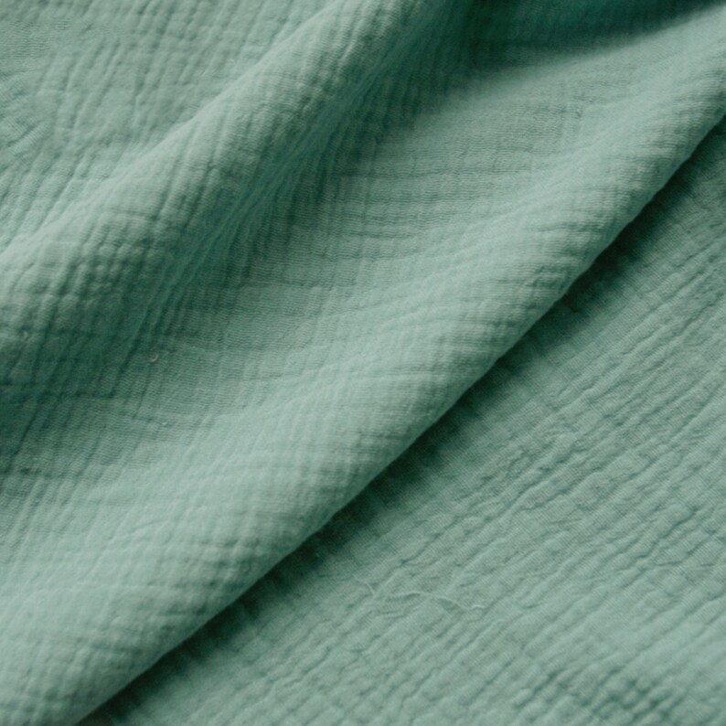 May include: A close-up shot of a folded piece of soft, textured fabric in a muted teal colour. The fabric has a crinkled, slightly puckered appearance, with visible diagonal and horizontal lines creating a grid-like pattern. The folds create shadows, adding depth.