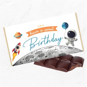 May include: A white chocolate bar with a birthday message that says "have a sweet birthday". The message is on a white background with a cartoon astronaut, rocket, and planet. The chocolate bar is partially eaten.