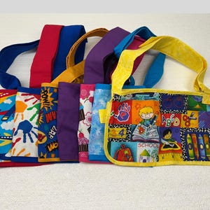 May include: A selection of brightly coloured fabric tote bags with diverse patterns. The bags feature straps in various colours, such as blue, red, yellow, and purple. The bag designs include school-themed graphics, handprints, and comic book-style text.