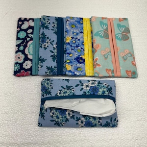 Könnte beinhalten: Eine Sammlung von Stoff-Taschentuchbox-Hüllen mit verschiedenen Blumen- und Schmetterlingsmustern. Die Hüllen zeigen Blau-, Gelb- und Rosatöne mit kontrastierenden Zierleisten. Eine Hülle ist geöffnet und zeigt weiße Taschentücher.