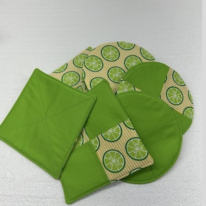 Puede incluir: Un conjunto de posavasos y agarraderas de tela hechos a mano. El conjunto incluye artículos cuadrados, redondos y de forma irregular. Las piezas lisas son de color verde brillante, mientras que otras presentan un estampado de rodajas de lima sobre un fondo a rayas crema y amarillo.