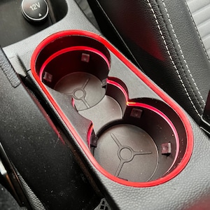 Op de afbeelding: Twee zwarte bekerhouders met een rode rand en een rood LED-licht rond de rand. De bekerhouders bevinden zich in de middenconsole van een auto.