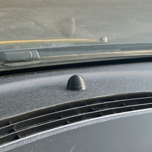May include: A small, black, dome-shaped object, possibly a car part, sits on a textured, dark dashboard. The object has a ridged surface and a vertical line down the center. The background includes a windshield and other interior car details.