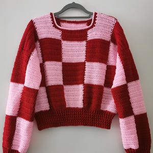 Könnte beinhalten: Ein handgehäkelter Pullover in einem rot- und hellrosa Schachbrettmuster. Der Pullover hat einen Rundhalsausschnitt und lange Ärmel. Das Garn scheint ein weiches Material mittlerer Stärke zu sein.