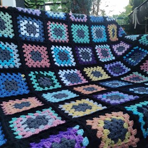 Könnte beinhalten: Eine schwarze Häkeldecke mit bunten Quadraten. Die Quadrate sind in einem Granny-Square-Muster gearbeitet und zeigen eine Vielzahl von Farben, darunter Rosa, Blau, Grün, Gelb und Lila.