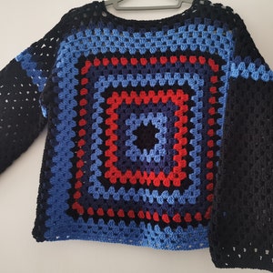 Op de afbeelding: Een gebreide trui met lange mouwen in zwart, blauw en rood. De trui heeft een groot, kleurrijk granny square-ontwerp in het midden.