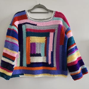 Può includere: Un maglione all'uncinetto fatto a mano e colorato con scollatura quadrata. Il design patchwork presenta vari colori tra cui blu, rosa, giallo, arancione e viola. Il maglione a maniche lunghe è appeso a una gruccia grigia.
