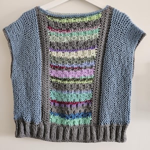 Könnte beinhalten: Ein handgestricktes ärmelloses Oberteil in Blau-, Grau- und mehrfarbigen Streifen. Das Oberteil hat einen gerippten Saum und ein Mittelteil mit strukturierten, horizontalen Streifen in verschiedenen Farben. Die Ärmel und der Rücken sind einfarbig hellblau.