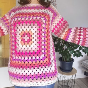 Könnte beinhalten: Ein rosa, weißer und orangefarbener Häkelpullover mit einem großen quadratischen Muster.