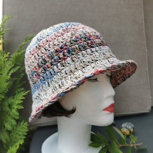 Op de afbeelding: Een gehaakte bucket hat met een meerkleurig gestreept patroon in tinten roze, blauw, wit en zwart. De hoed heeft een brede rand en is gemaakt met een textuur.