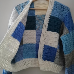 Könnte beinhalten: Eine gehäkelte Strickjacke mit Patchwork-Muster in Blautönen und Weiß. Die Strickjacke hat eine lockere Passform und einen langen, offenen Vorderteil.