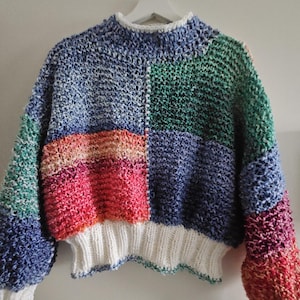 Może przedstawiać: Ręcznie robiony sweter z blokami kolorów i wysokim dekoltem. Sweter ma wzór patchworkowy z niebieskimi, zielonymi, różowymi i czerwonymi sekcjami. Mankiety i dół są białe. Sweter jest wykonany z grubego materiału.