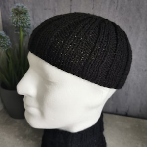 Könnte beinhalten: Eine schwarze Strickmütze mit Rippenmuster. Die Mütze wird auf einem weißen Schaufensterkopf präsentiert, um ihre Form und Textur zu zeigen. Die Mütze ist aus einem dunklen, möglicherweise wollenen Material gefertigt.