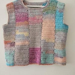 Könnte beinhalten: Eine handgefertigte Häkelweste im Patchwork-Design. Die Weste hat eine Vielzahl von Pastellfarben, darunter Rosa, Blau, Gelb und Grau. Das ärmellose Oberteil wird auf einem weißen Kleiderbügel vor einem weißen Hintergrund präsentiert. Die Weste ist aus weichem Garn gefertigt.