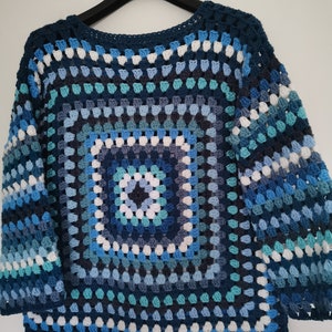 Könnte beinhalten: Ein blauer und weißer Häkelpullover mit einem quadratischen Muster in der Mitte. Der Pullover hat eine lockere Passform und ist aus verschiedenen Blautönen gefertigt.