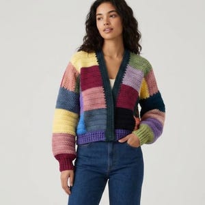 Può includere: Un cardigan all'uncinetto a maniche lunghe con un design patchwork a blocchi di filato giallo, rosa, bordeaux, blu, viola, verde e nero. Il capo presenta uno scollo a V, un bordo a coste verde scuro lungo l'apertura e polsini a coste.