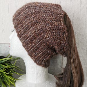 Könnte beinhalten: Ein brauner gehäkelter Ponytail-Beanie-Hut. Der Hut besteht aus einer Mischung aus braunem und beigem Garn mit strukturiertem Aussehen. Der Beanie ist für einen Pferdeschwanz konzipiert, mit einer Öffnung auf der Rückseite. Das Haar ist dunkelbraun.