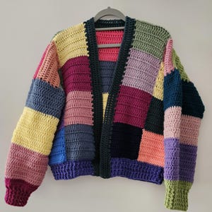 Könnte beinhalten: Ein handgefertigter Patchwork-Häkelcardigan in verschiedenen Farben, darunter Gelb, Rosa, Lila und Blau. Der offene Pullover hängt an einem grauen Kleiderbügel vor einem schlichten weißen Hintergrund. Die Ärmel und der Körper bestehen aus rechteckigen Blöcken.