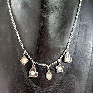Peut inclure: Un collier à chaîne argentée avec cinq breloques pendantes, chacune contenant une petite perle ronde blanc cassé. Les breloques ont la forme d'un losange, d'un cœur, d'une goutte d'eau, d'une étoile et d'un second cœur, suspendues à la chaîne.