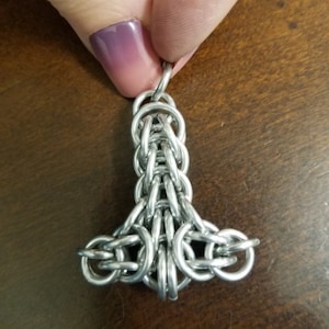 Puede incluir: Un colgante de metal plateado en forma de Mjolnir, el martillo de Thor, un dios nórdico. El colgante está hecho de anillos de metal entrelazados.