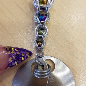 May include: A silver chain with colorful beads and a silver disc pendant. The chain is made of small, interconnected rings. The beads are iridescent and have a rainbow effect. The disc pendant is smooth and has a slightly textured surface.