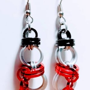 May include: A pair of dangle earrings with silver, black, and red metal rings. The earrings have a silver hook and a chain with a small red ring at the bottom.