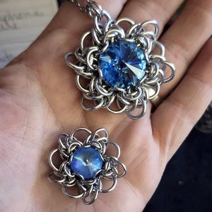 Könnte beinhalten: Ein silberfarbenes Kettenanhänger-Halsketten- und passendes Ohrstecker-Set. Jedes Stück hat einen großen, facettierten, leuchtend blauen Kristall in der Mitte, umgeben von einem Kettenhemd-Blumenmuster. Der Schmuck wird auf einer Hand präsentiert.