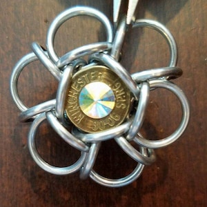 May include: A silver metal flower charm with a brass bullet casing in the centre. The bullet casing has a clear iridescent stone in the centre and the text "WINCHESTER 30-06" printed around the stone.