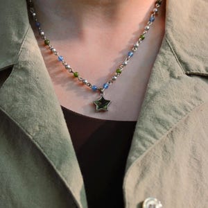 May include: A silver necklace with a star pendant. The necklace features small, colorful beads in blue, green, and silver. The star pendant is a five-pointed star with a dark green center. The necklace is worn over a dark brown top and a light green jacket.
