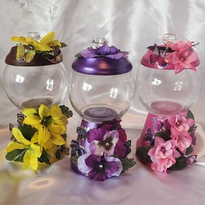 May include: Three glass candy jars with decorative lids and bases. The jars are clear glass and the lids and bases are painted in different colors. The lids are decorated with flowers and butterflies. The bases are decorated with flowers and butterflies. The jars are on a white surface.