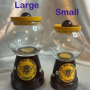 May include: Two glass candy jars with yellow lids and brown bases. The bases are decorated with a bee and the words "Kind. Wild. Humble. Loving. Yourself. Happy."