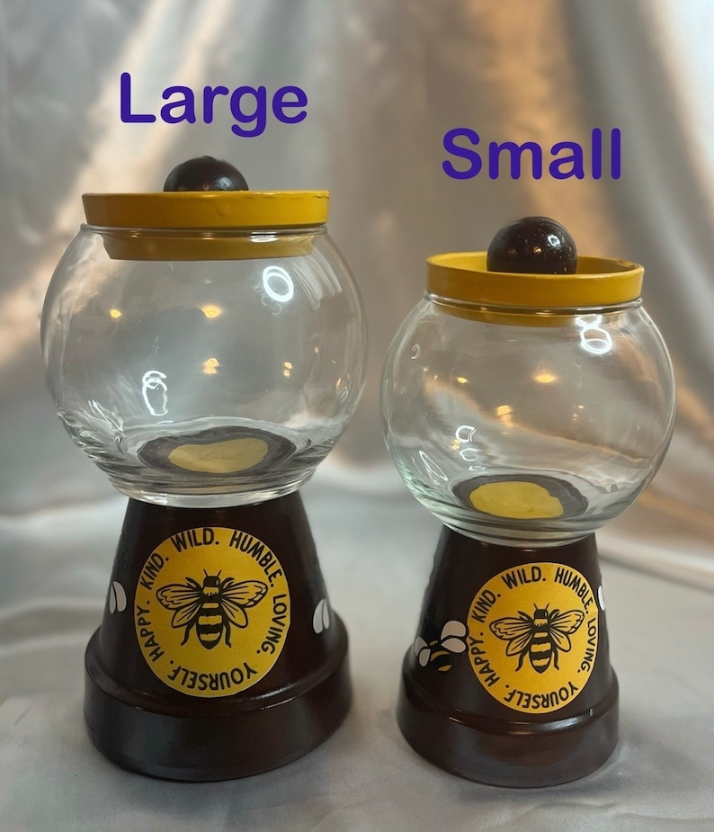 May include: Two glass candy jars with yellow lids and brown bases. The bases are decorated with a bee and the words "Kind. Wild. Humble. Loving. Yourself. Happy."