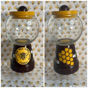 May include: A glass candy jar with a yellow lid and a brown base decorated with a honeycomb pattern and a bee. The base also has a yellow circle with the text "Happy. Kind. Wild. Humble. Loving. Yourself." around a bee.