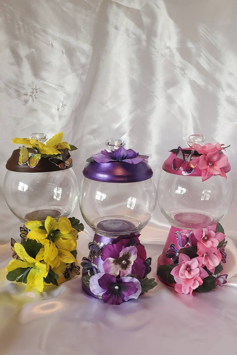 May include: Three glass candy jars with decorative lids and bases. The jars are clear glass and the lids and bases are painted in different colors. The lids are decorated with flowers and butterflies. The bases are decorated with flowers and butterflies. The jars are on a white surface.