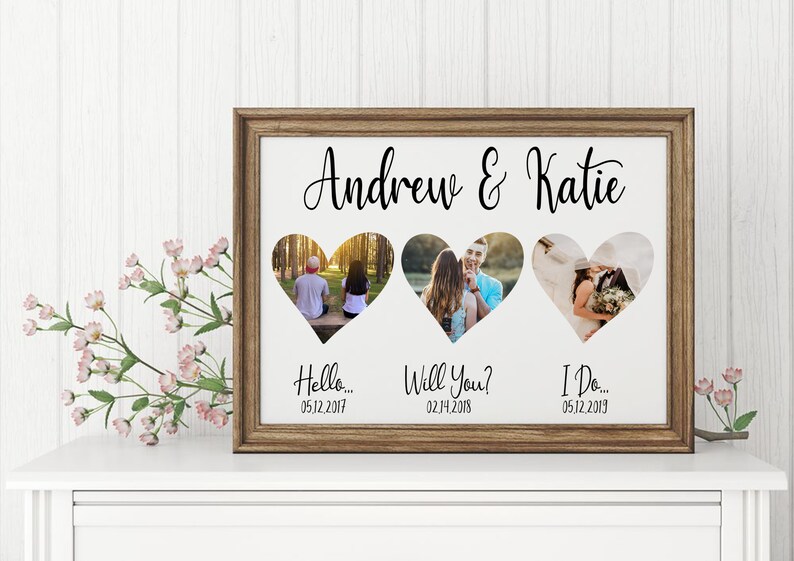 K&ouml;nnte beinhalten: Ein gerahmtes Fotocollage mit drei herzf&ouml;rmigen Ausschnitten, die jeweils ein anderes Foto zeigen. Der Rahmen ist braun, und der Text "Andrew & Katie" steht &uuml;ber den Fotos. Darunter stehen die S&auml;tze "Hello...", "Will You?" und "I Do..." mit Daten.