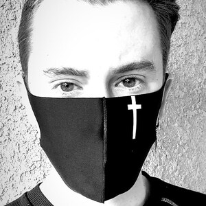 May include: A person wearing a black face mask with a white cross design. The mask covers the person's nose and mouth. The person has short, dark hair and is looking directly at the camera.