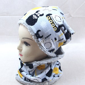 Könnte beinhalten: Eine graue und weiße Strickmütze und ein passender Halsschmuck mit einem schwarzen und gelben Druck eines Wikingerschiffs und eines Bären mit Helm. Mütze und Halsschmuck sind mit einem weichen grauen Fleece gefüttert.